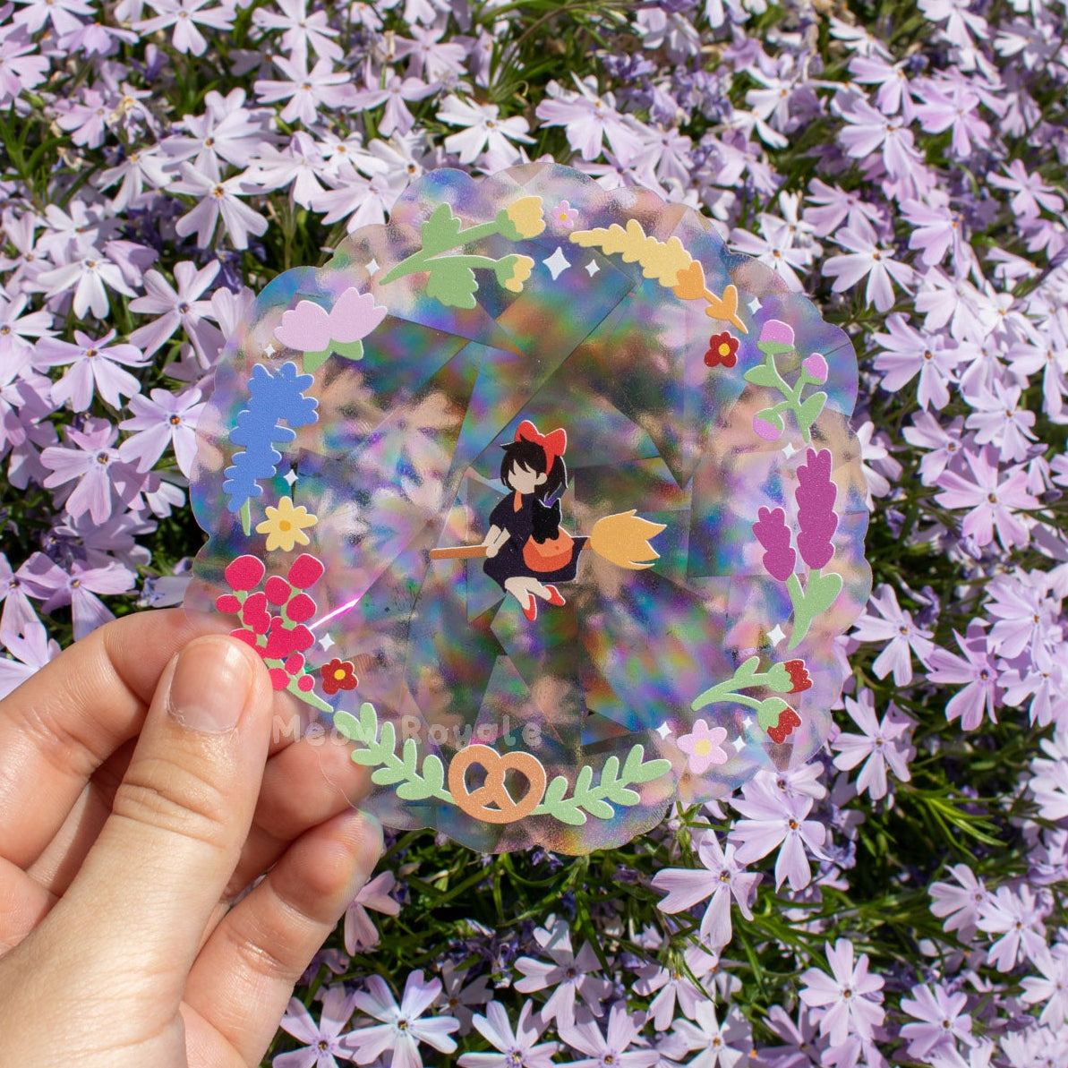 Hand holding a colorful sticker with a character surrounded by purple flowers