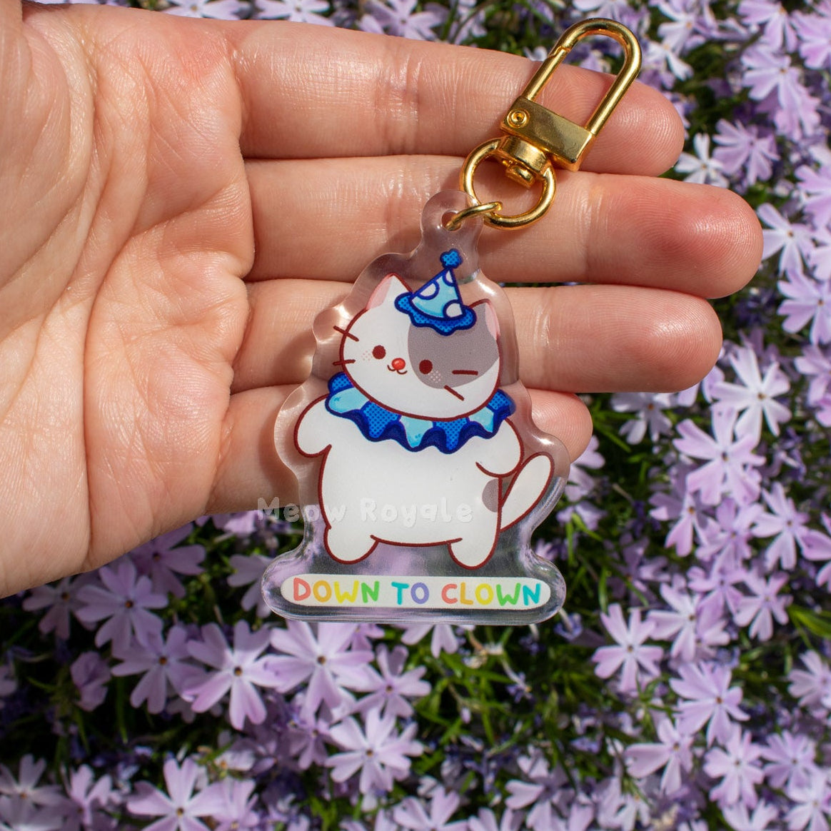 Hand holding a clear acrylic keychain with cartoon character and 'Down to Clown' text against a background of purple flowers.